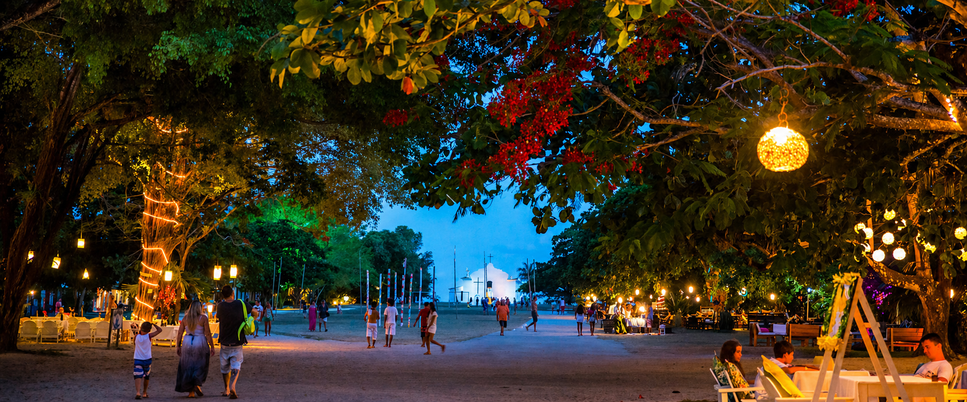 Vista noturna do Quadrado de Trancoso, Bahia, com árvores iluminadas por lanternas, pessoas caminhando e a igrejinha branca ao fundo sob um céu azul profundo.