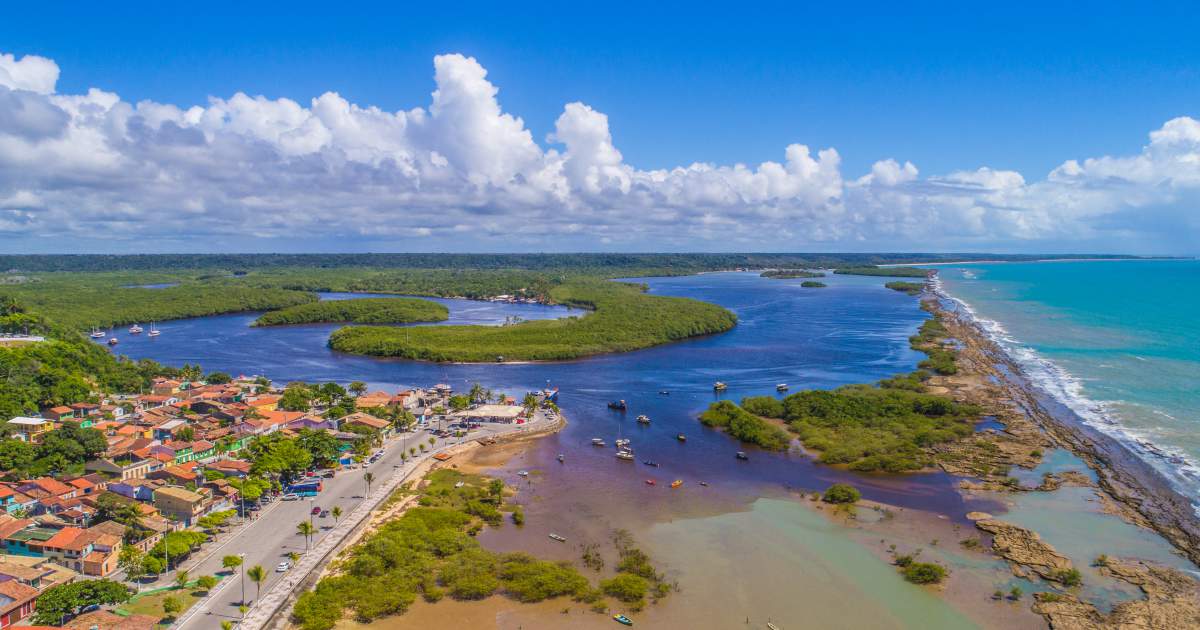 Vista aérea do encontro do rio com o mar em Santa Cruz Cabrália, Bahia, mostrando a orla, áreas verdes de manguezal e o oceano azul sob um céu com nuvens brancas.