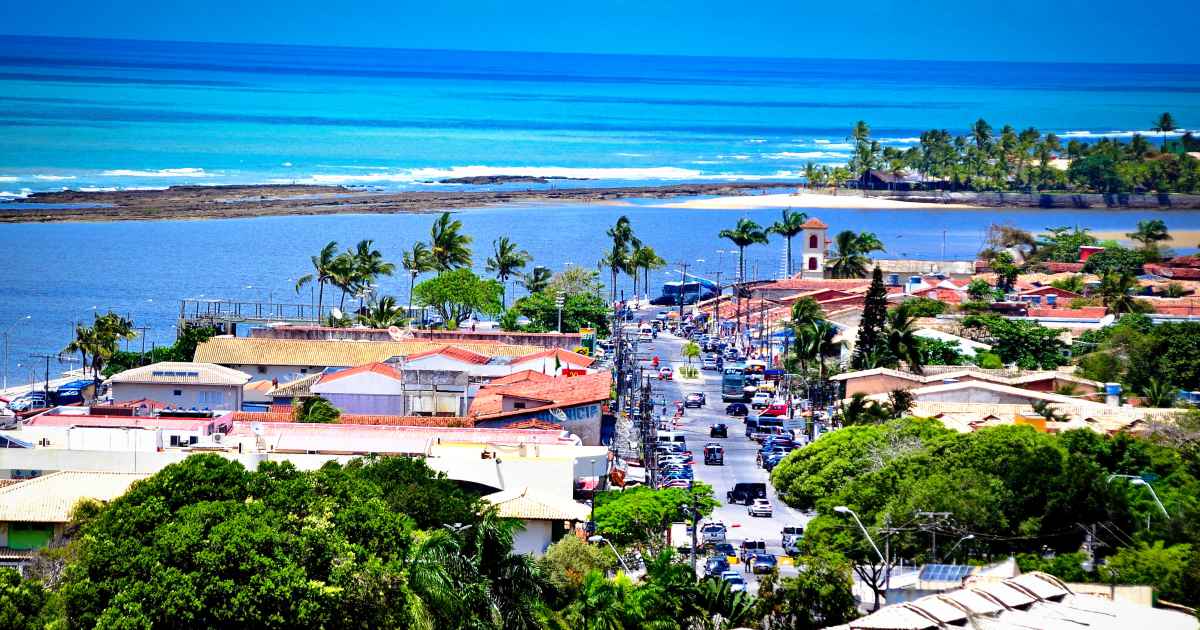 Vista aérea da cidade de Porto Seguro com casas de telhado cerâmico, ruas arborizadas e o mar azul claro com recifes de coral ao fundo sob um céu limpo.